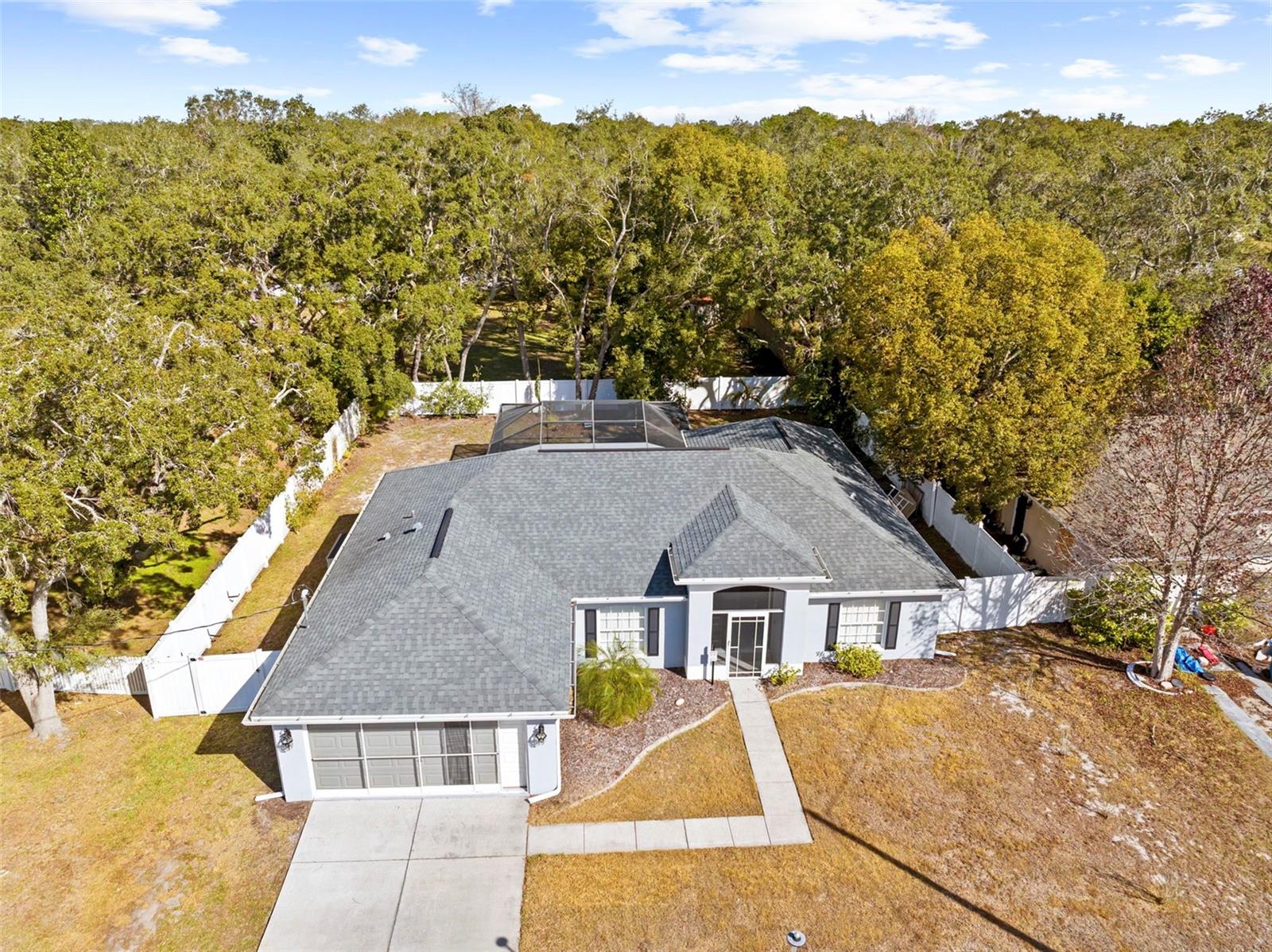 Aerial view of a single-story house with a gray roof, surrounded by trees and a white fence. The yard is mostly grass with a concrete walkway leading to the front door.