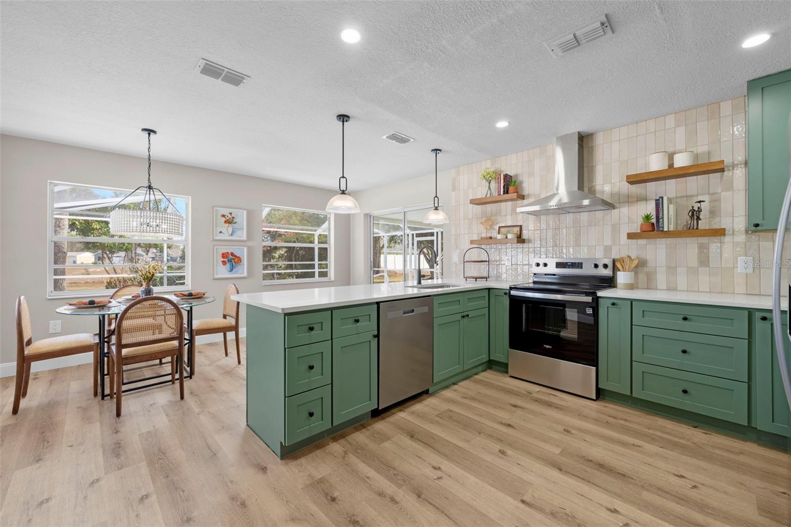 Modern kitchen featuring green cabinetry, stainless steel appliances, and a spacious island with a white countertop. A dining table with four chairs is situated near large windows, adorned with decorative art and houseplants.