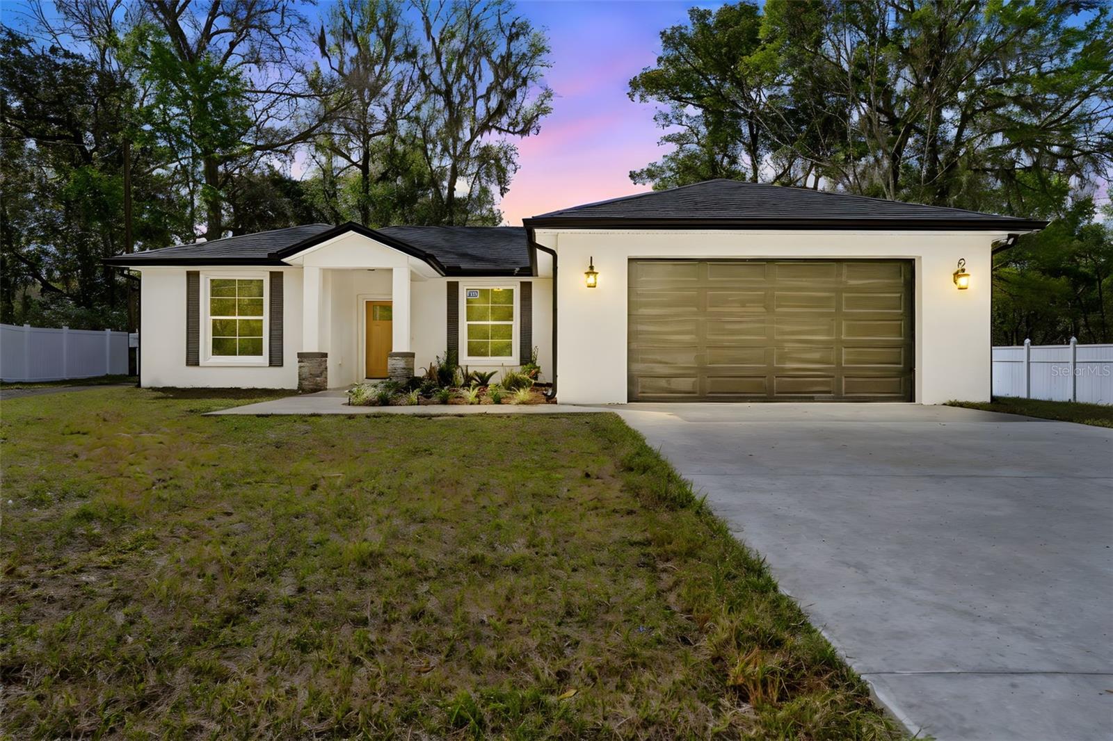 Exterior view of a modern single-family home featuring a light-colored facade, a sloped roof, and a two-car garage, surrounded by lush greenery and a concrete driveway.